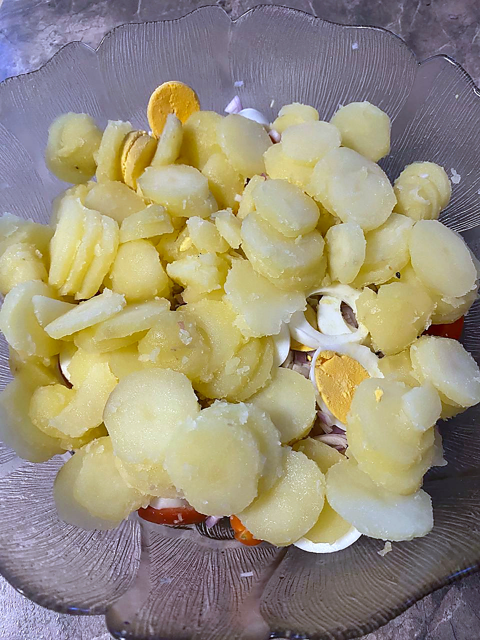 Sliced boiled potatoes added over eggs, red onion and cherry tomatoes in a glass mixing bowl.